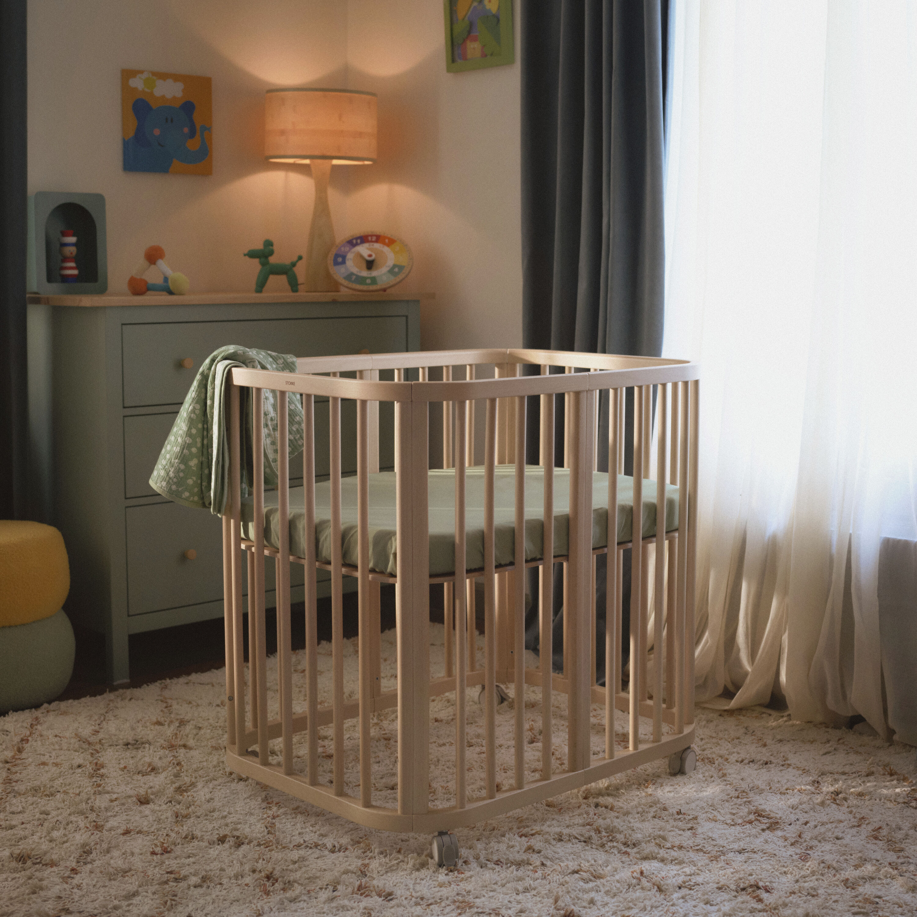 A lifestyle shot of the Stokke Nighti™ Mini Newborn Cot in a calm, simply arranged nursery, with clear space around the cot and a changing dresser visible in the background, conveying warmth and practicality.